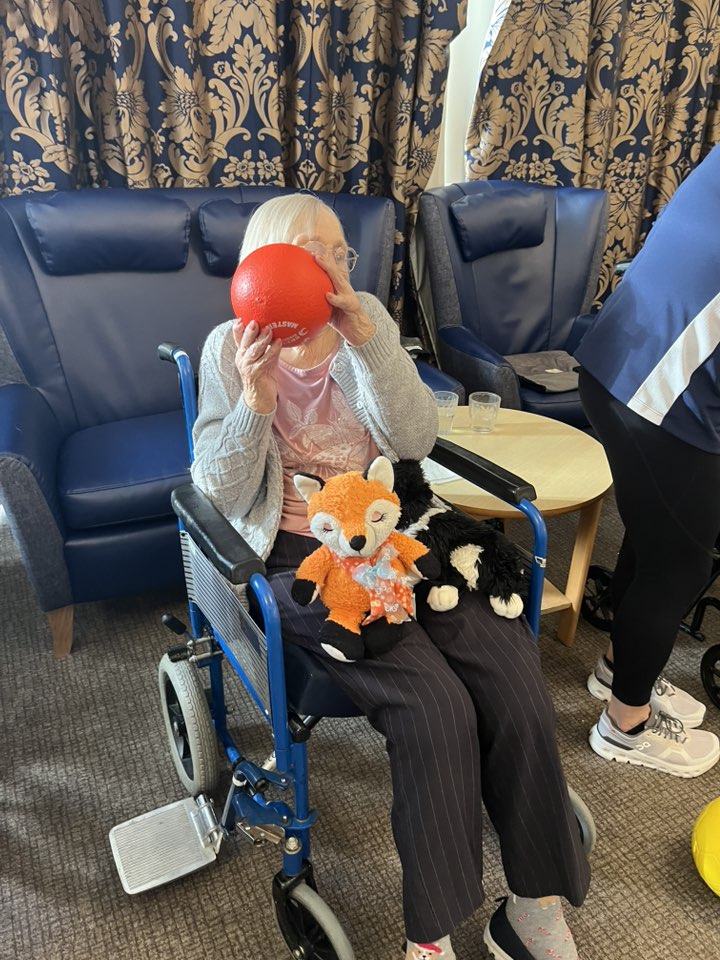 Elderly woman in a wheelchair holding a red ball with stuffed toys on her lap.