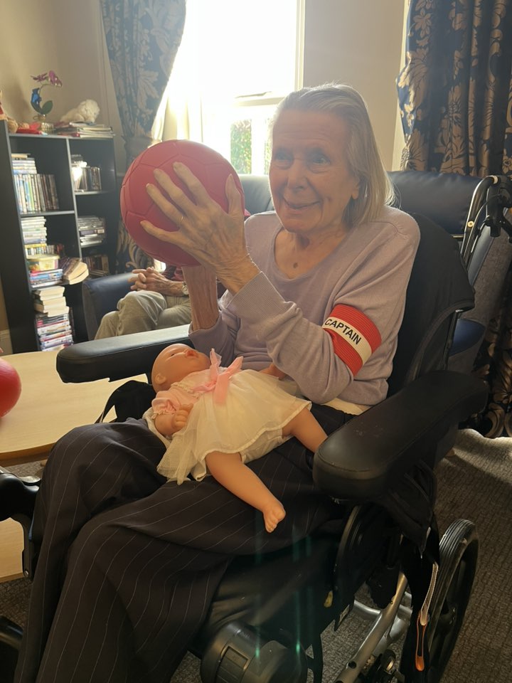 Elderly woman in a wheelchair holding a red ball and a doll in a bright living room.