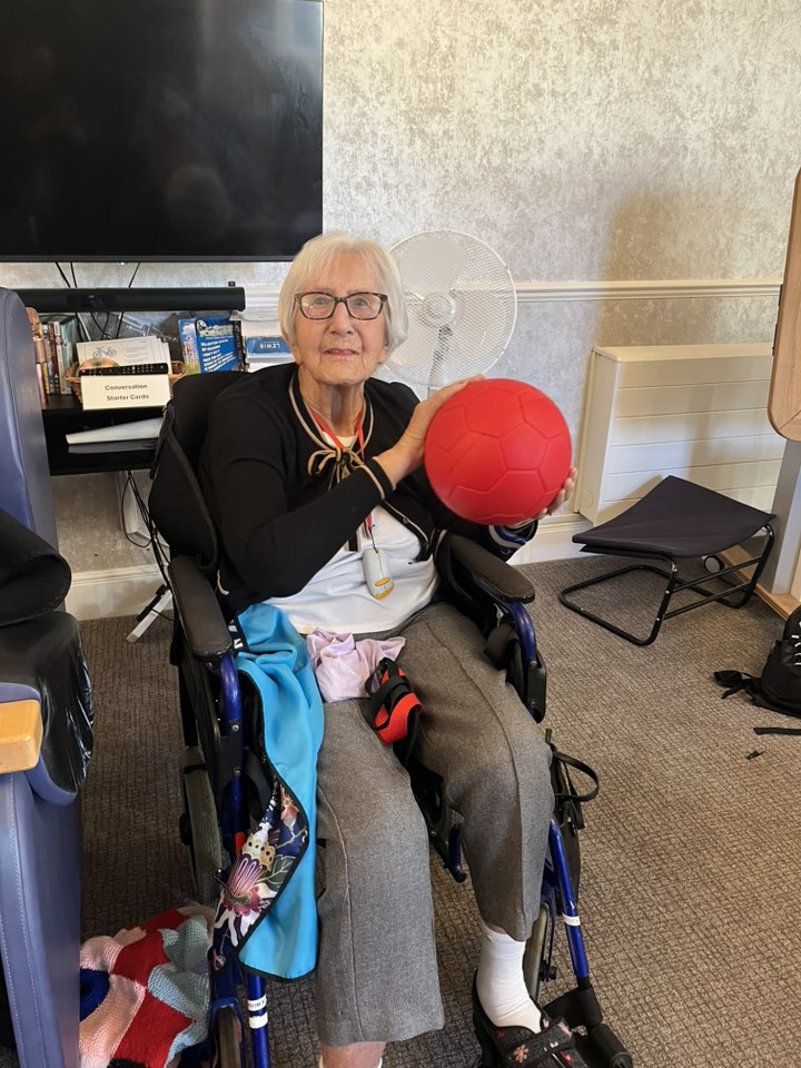 Elderly woman sitting in a wheelchair holding a red ball and looking at the camera.