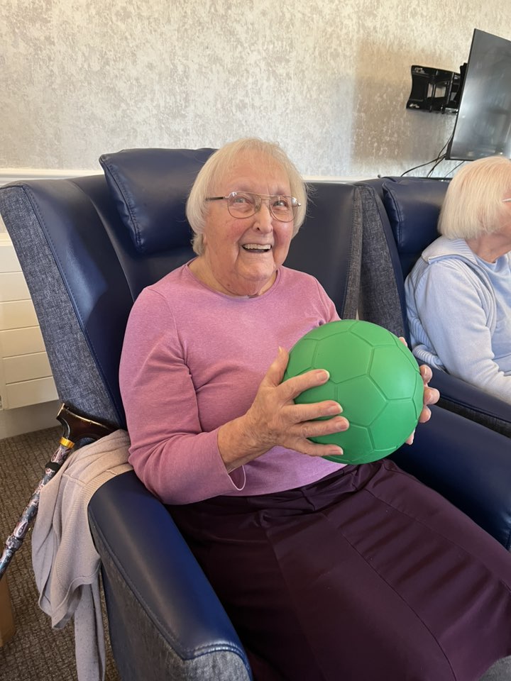 Elderly woman in a chair holding a green ball and smiling during a group activity.