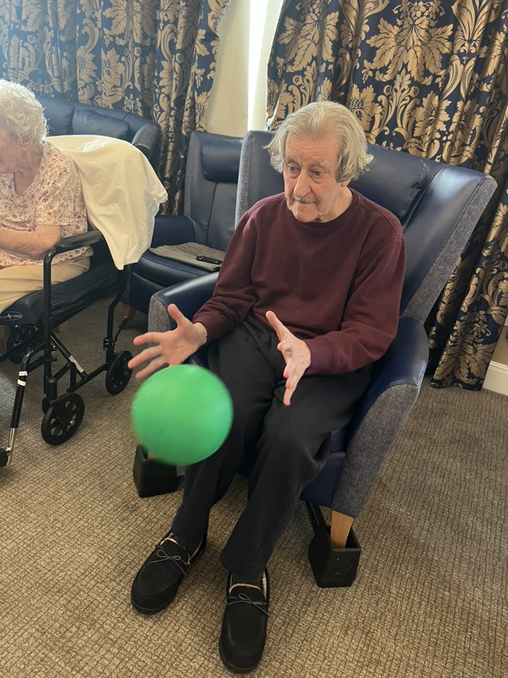 Elderly man seated, tossing a green ball during an activity.