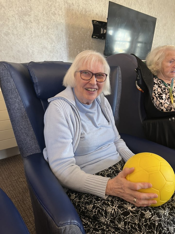 Smiling elderly woman seated in an armchair holding a yellow ball.