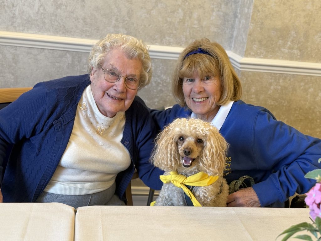 Two elderly women smiling with a small dog wearing a yellow bandana.
