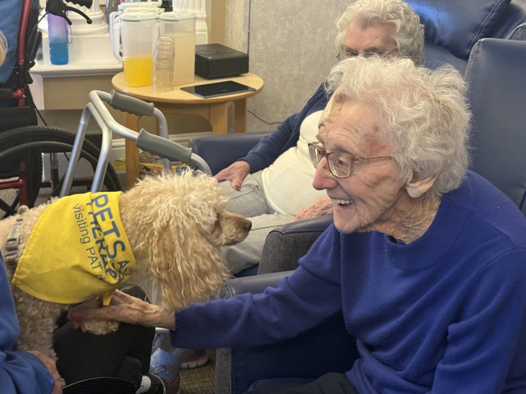 Elderly woman smiling and petting a small dog during a visit.