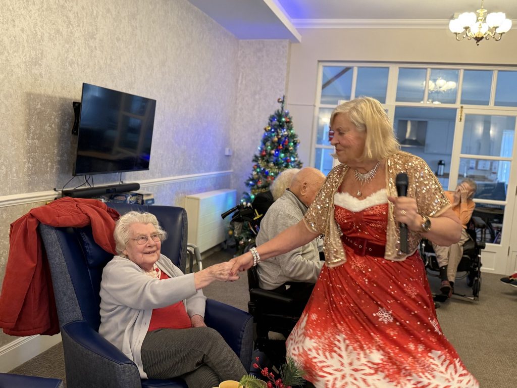 Woman in festive red dress holding microphone, dancing with seated elderly woman by a Christmas tree.