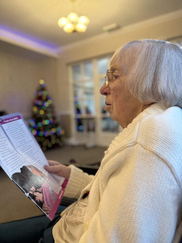 Elderly woman reading a booklet in a lounge with a decorated Christmas tree in the background.