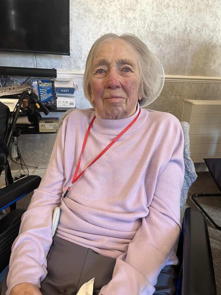 Elderly woman in a pink sweater sitting in a chair, looking at the camera.