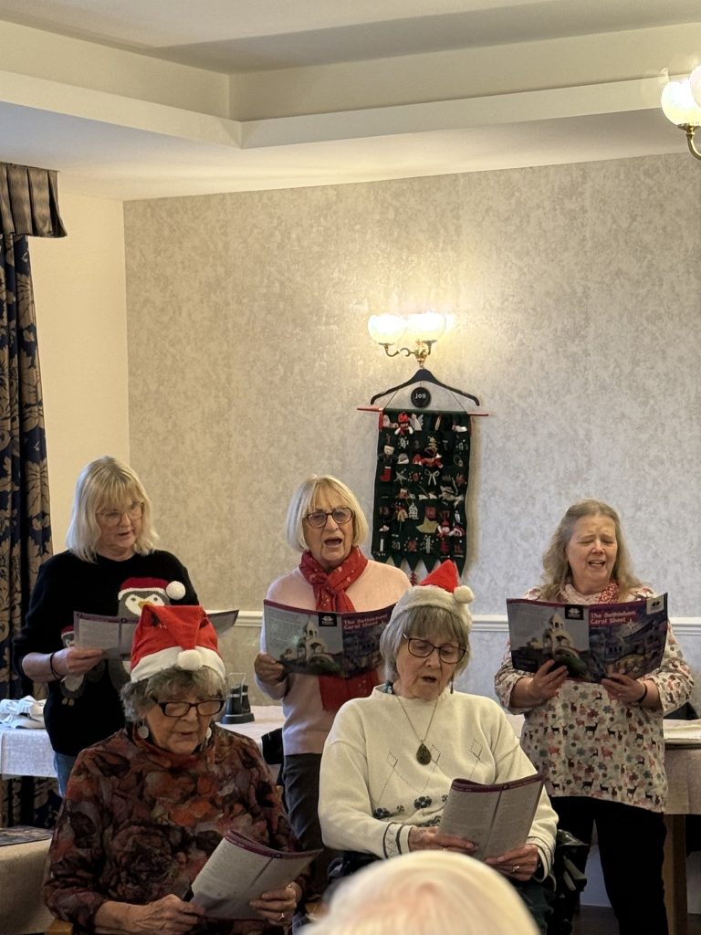 Group of women wearing Santa hats singing from song sheets in a holiday gathering.