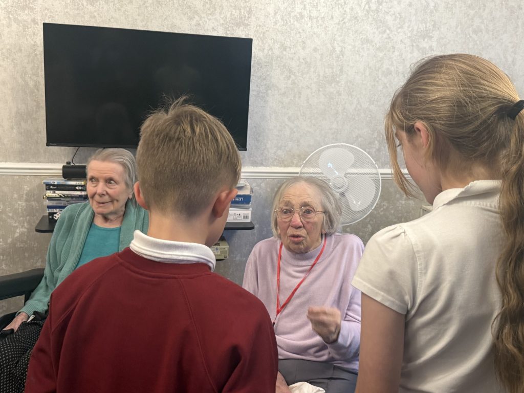 Elderly women chatting with two schoolchildren in a care home lounge.