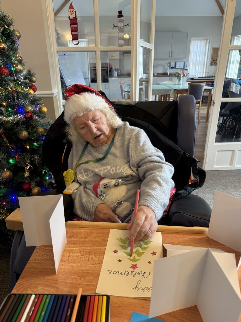 Elderly woman in a Santa hat coloring a handmade Christmas card at a table.