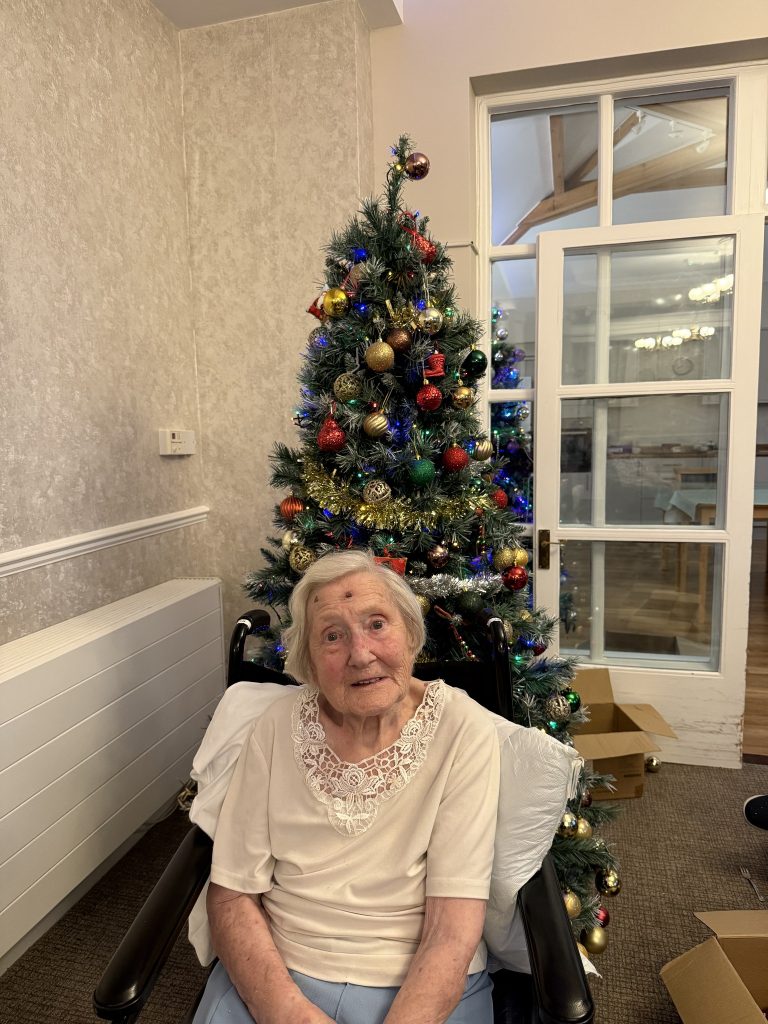 Elderly woman in a wheelchair sitting in front of a decorated Christmas tree.
