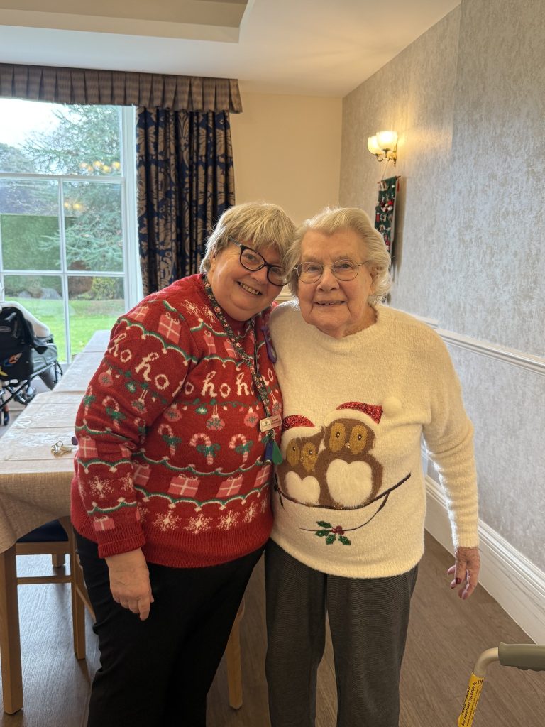 Two women smiling and standing close together in festive Christmas sweaters indoors.