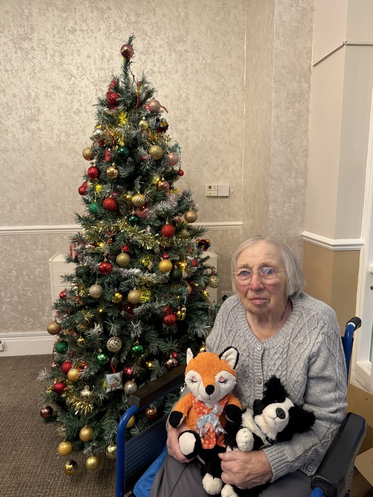 Elderly woman holding stuffed toys beside a decorated Christmas tree.