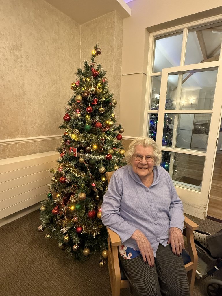 Smiling elderly woman sitting next to a lit Christmas tree in a cozy room.