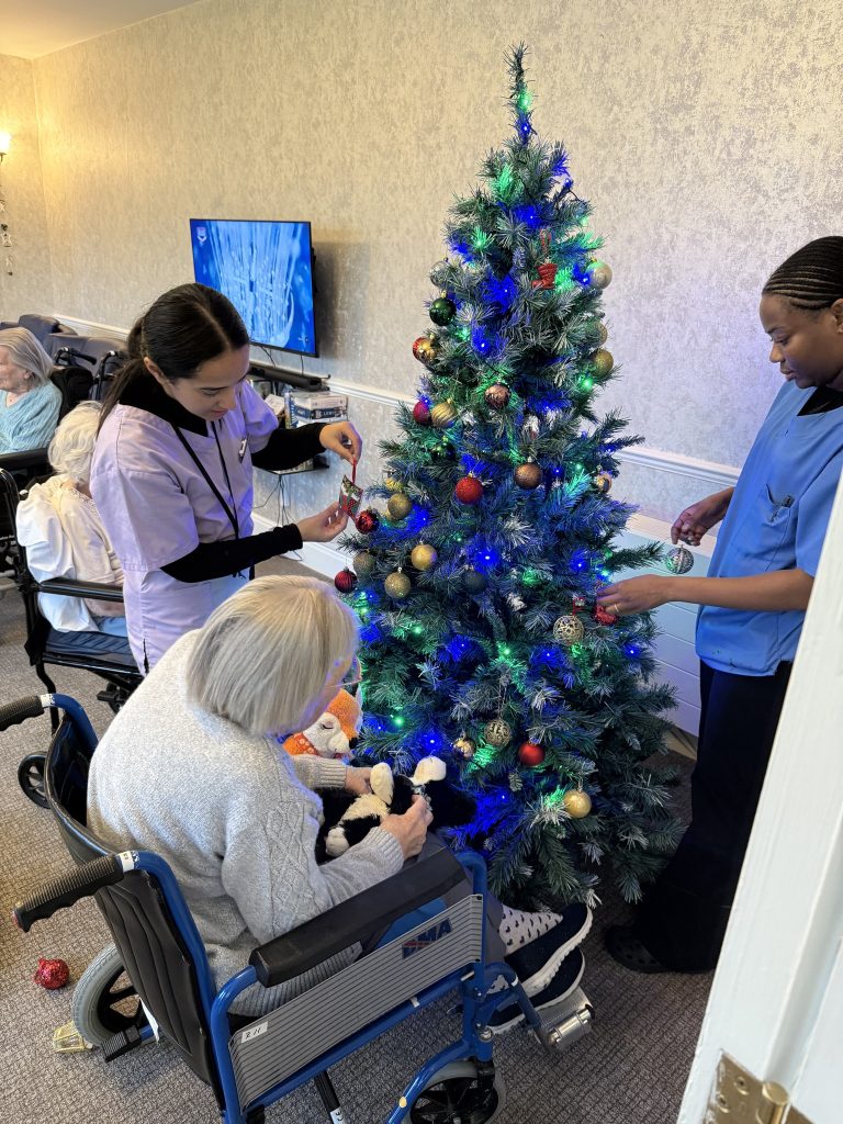 Staff and an elderly resident in a wheelchair decorating a lit Christmas tree together.