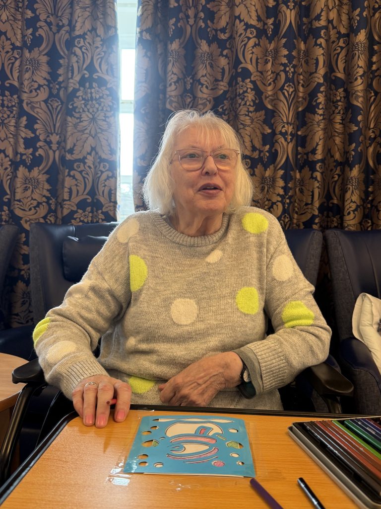Elderly woman sitting at a table using a stencil and colored pencils for a craft activity.