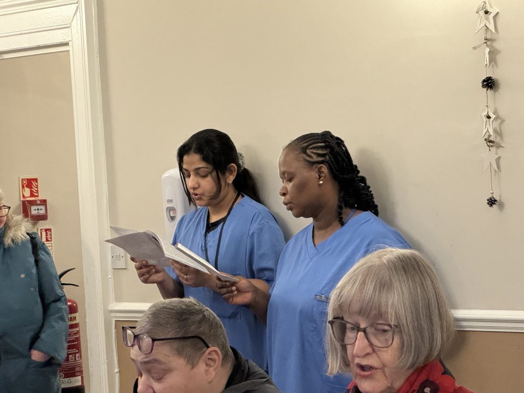 Two caregivers in blue uniforms read from papers while residents sit nearby.