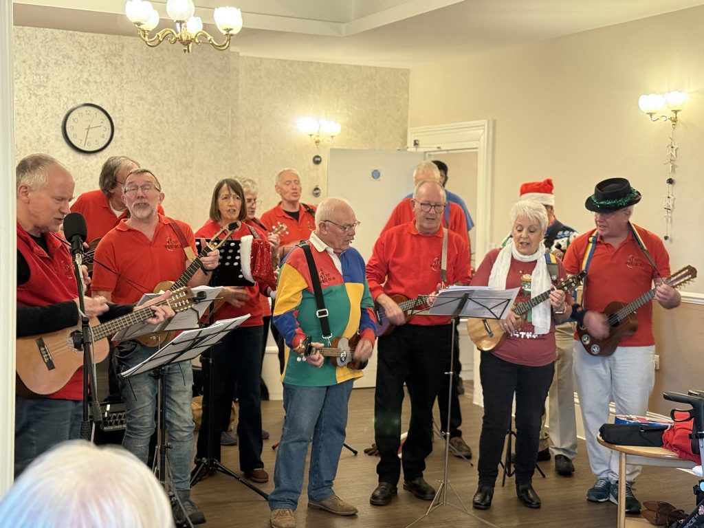 Group of musicians in festive clothing playing instruments and singing in a community room.