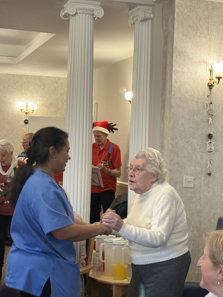 Care worker holding hands and dancing with an elderly woman while musicians perform in the background.