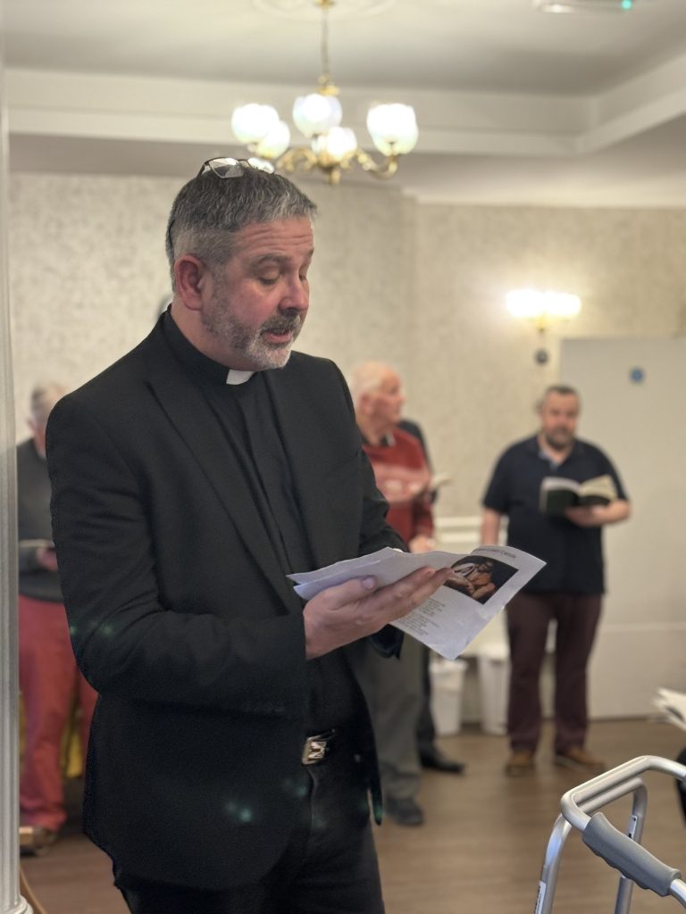 Priest reading from a booklet while a small group stands behind him holding books.