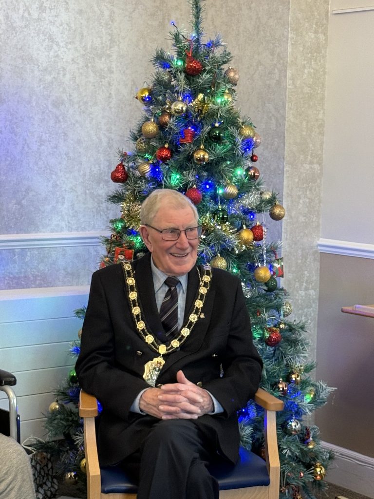 Elderly man in formal attire sitting and smiling in front of a decorated Christmas tree.