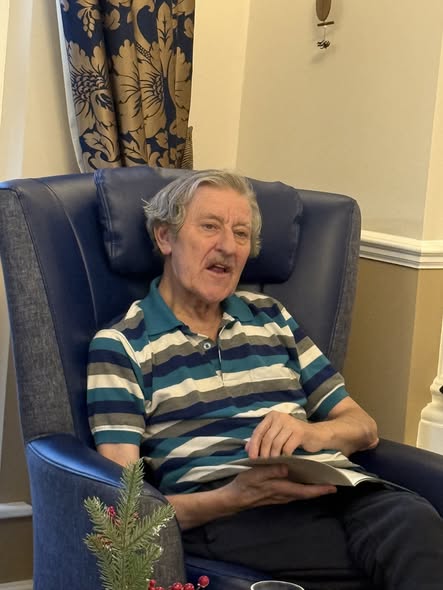 Elderly man sitting in an armchair, reading aloud from a booklet.