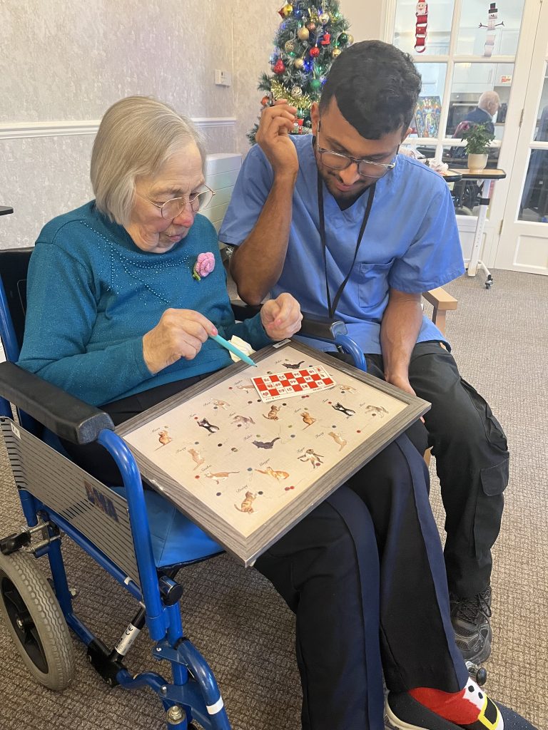 Elderly woman in a wheelchair and caregiver playing a board game together near a Christmas tree.