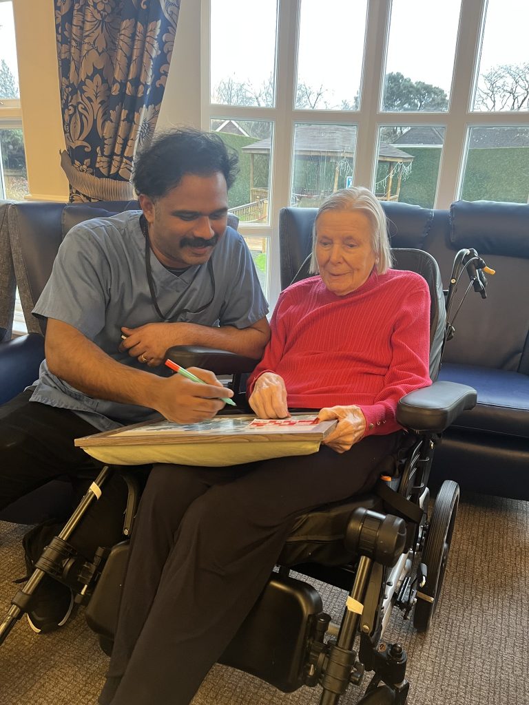 Caregiver assisting an elderly woman in a wheelchair with a tabletop game by a window.