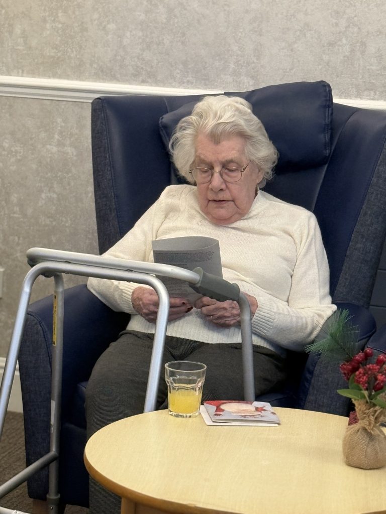 Elderly woman seated in a chair reading a leaflet, with a drink on a table beside her.