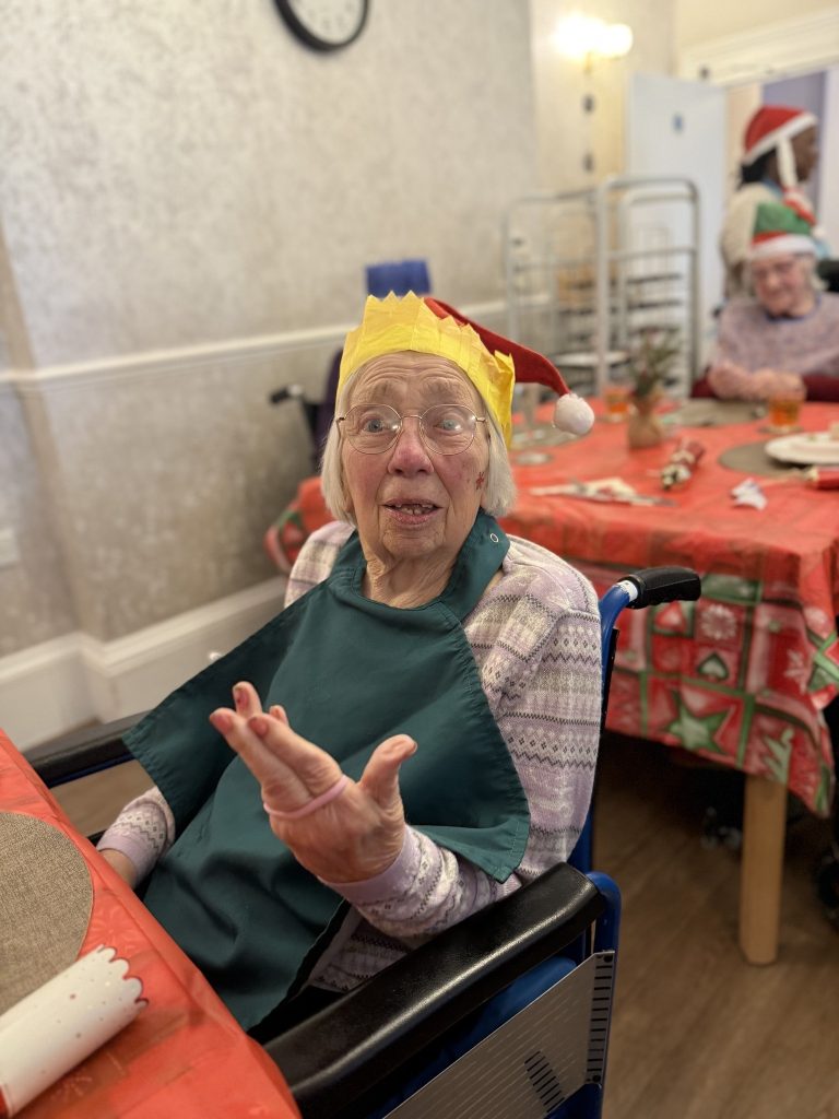Elderly man in a paper crown holding a glass of wine, smiling beside a caregiver in festive clothing at a Christmas meal.