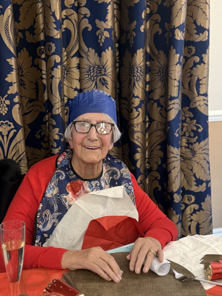 Elderly woman smiling at a festive table, wearing a paper crown and bib during a Christmas meal.