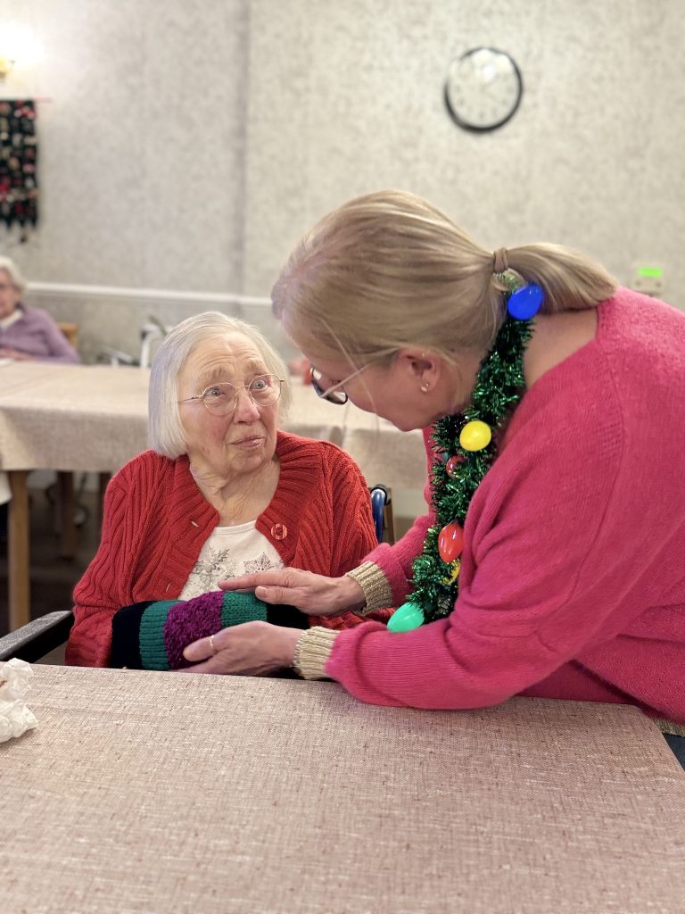 Caregiver helping an elderly woman try on a colorful knitted item at a table.