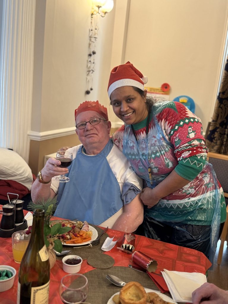 Elderly man in a paper crown holding a glass of wine, smiling beside a caregiver in festive clothing at a Christmas meal.