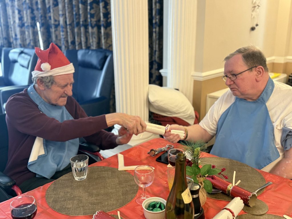 Two elderly men in a care home pulling Christmas crackers at a festive dining table.
