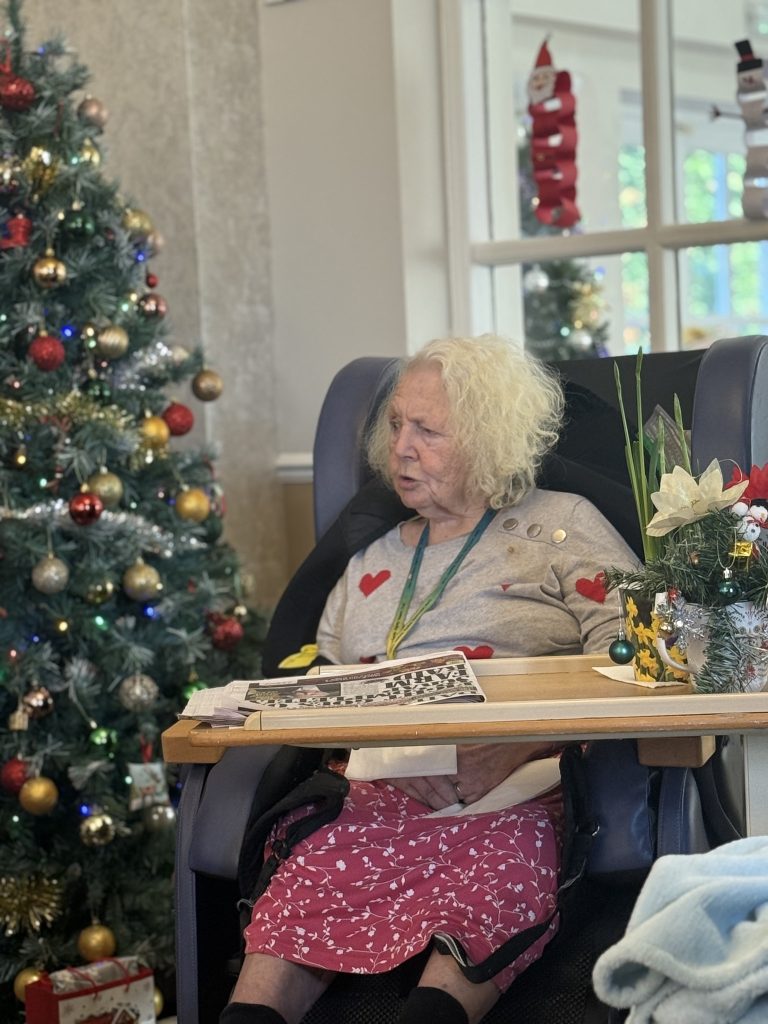 Elderly woman sitting by a decorated Christmas tree with a newspaper on her lap.