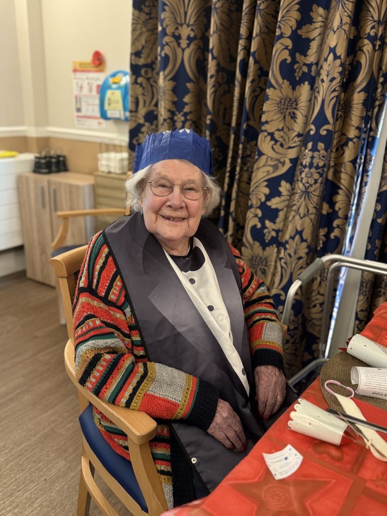 Smiling elderly woman wearing a blue paper crown and colorful cardigan, seated at a Christmas-themed table.