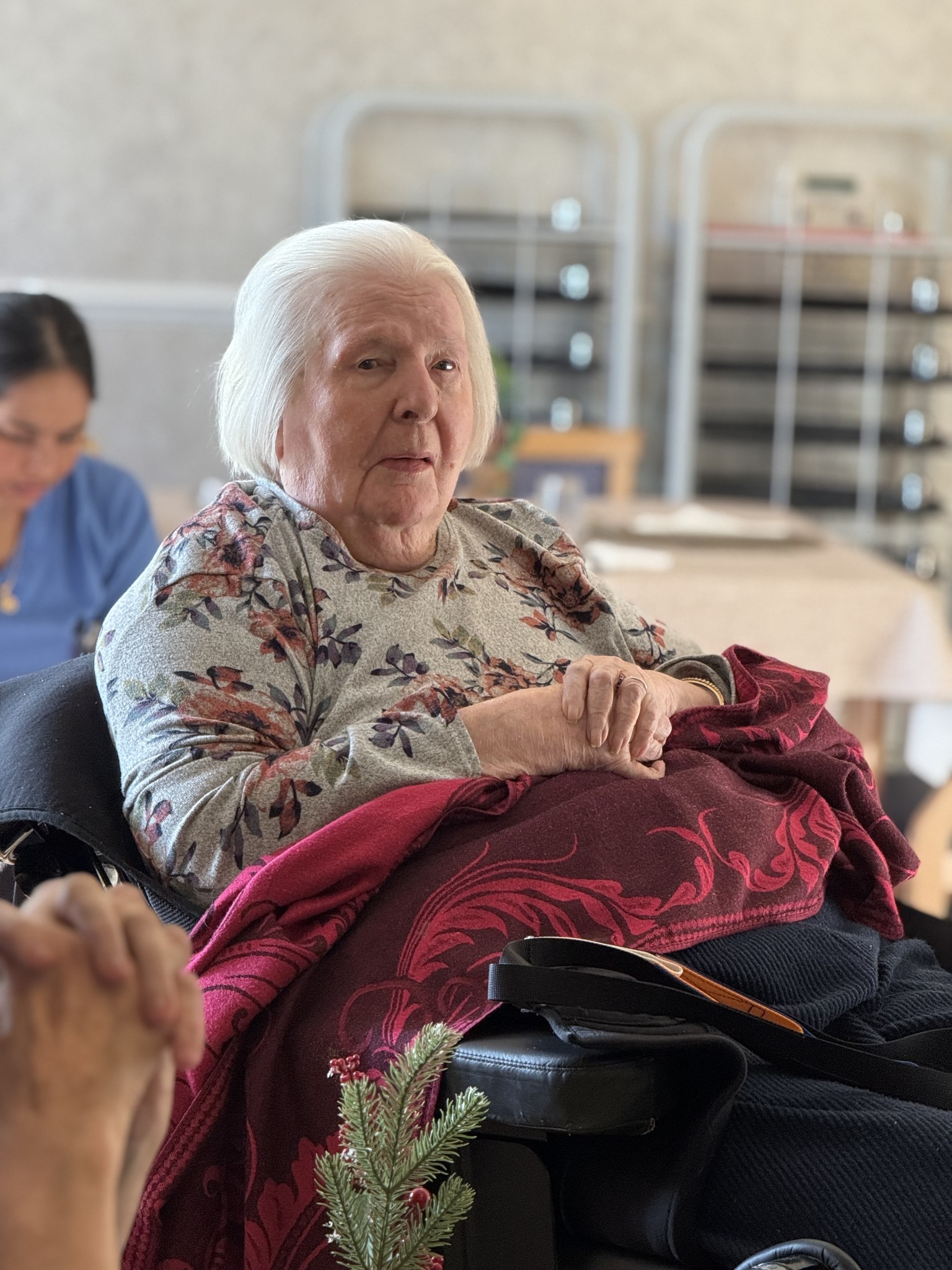 Elderly woman sitting in a chair with a blanket over her lap in a care home setting.