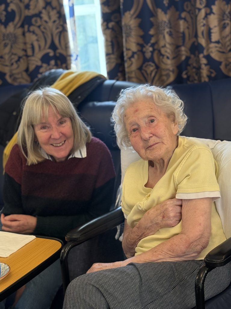 Two elderly women sitting together, smiling and looking at the camera in a cozy room.