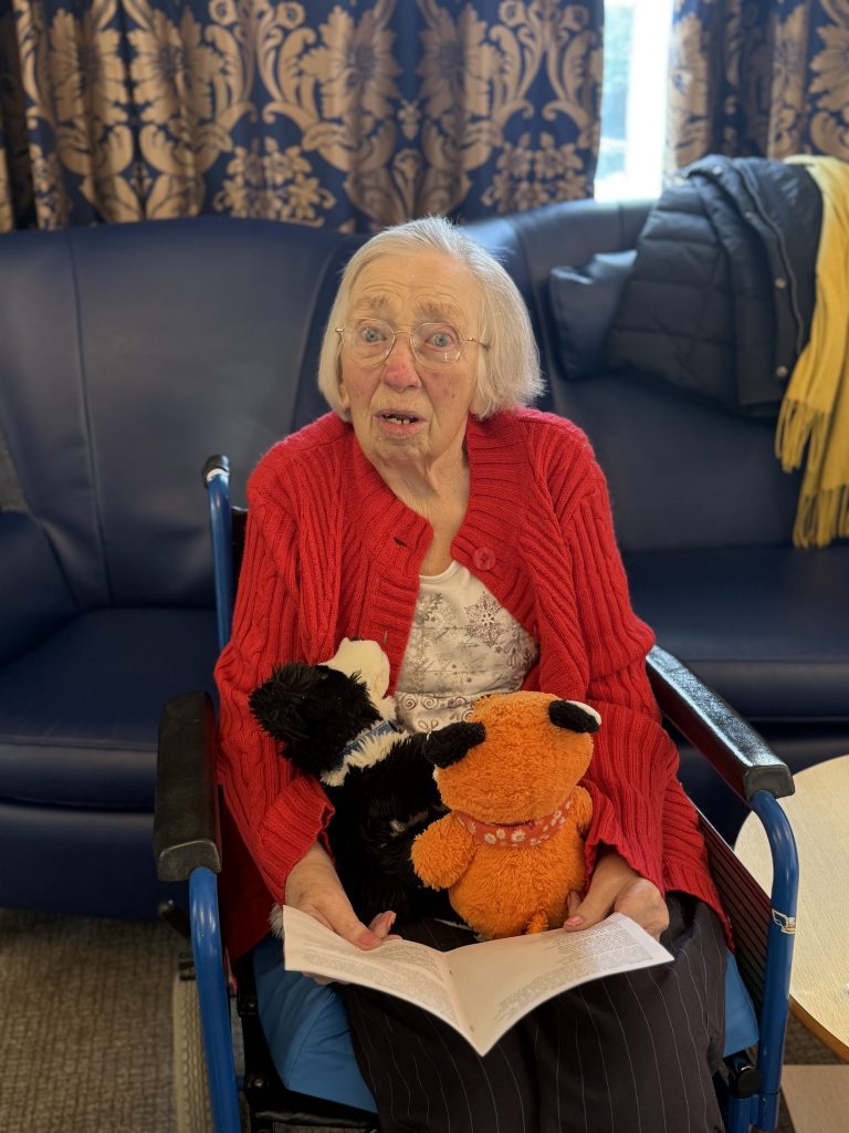 Elderly woman in a wheelchair holding soft toys and reading a card, looking surprised.