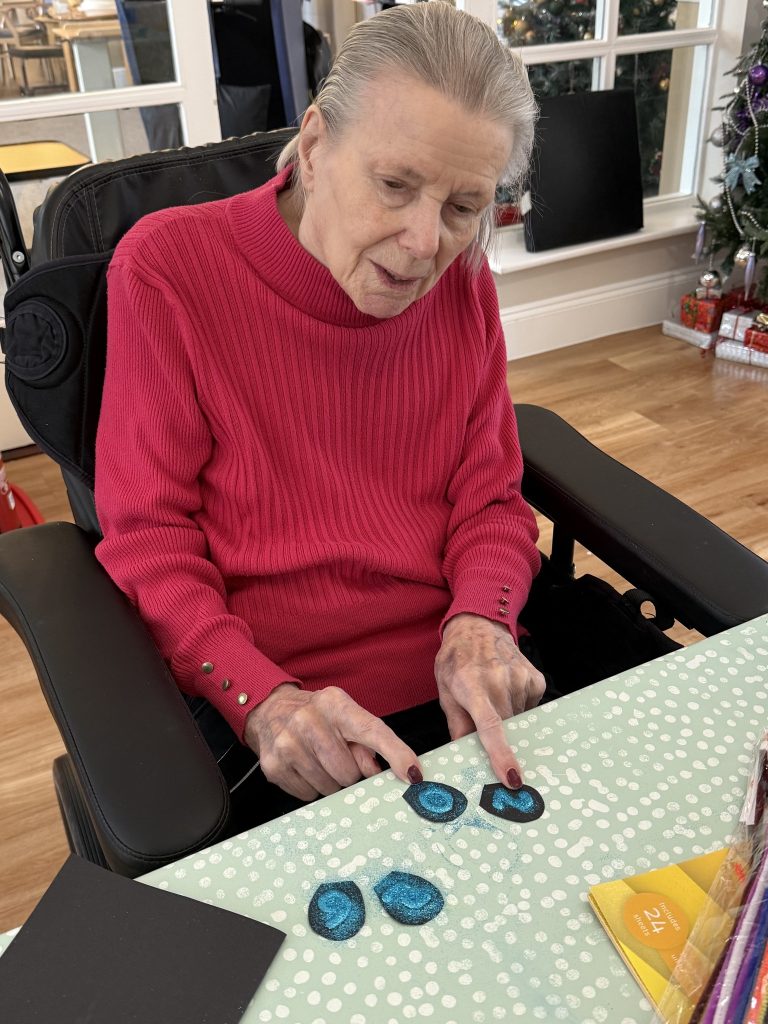 Elderly woman arranging glittery shapes on a table during a craft activity.