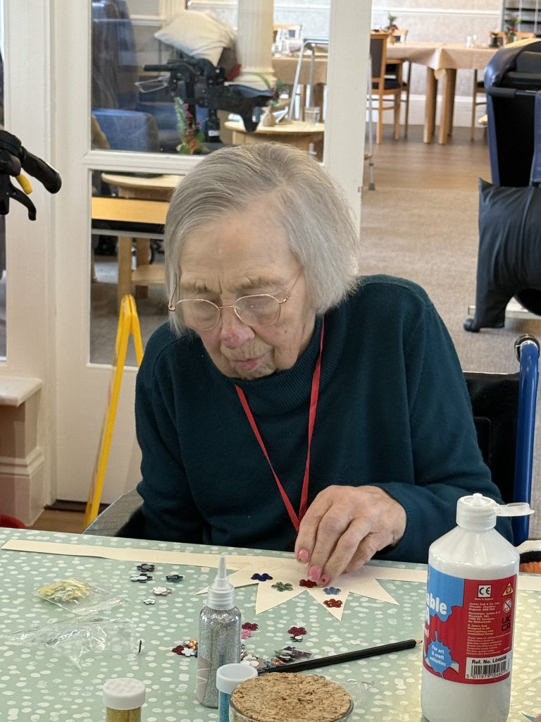 Elderly woman carefully decorating a paper star with sequins at a craft table.