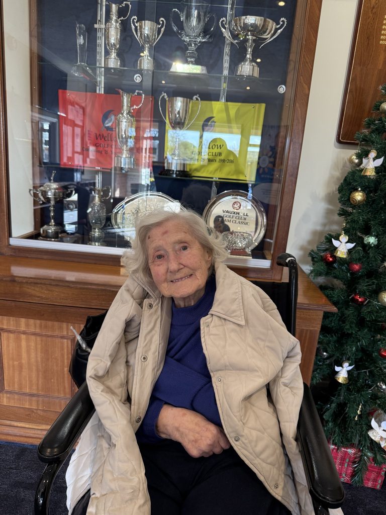 Elderly woman smiling in a wheelchair in front of a trophy display and Christmas tree.