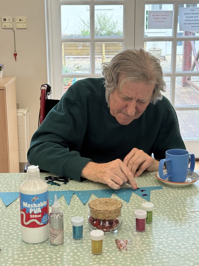 Elderly man concentrating while decorating bunting with glitter and craft supplies.