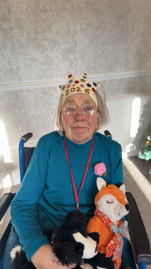 Elderly woman wearing a paper crown holding soft toys while seated in a wheelchair.