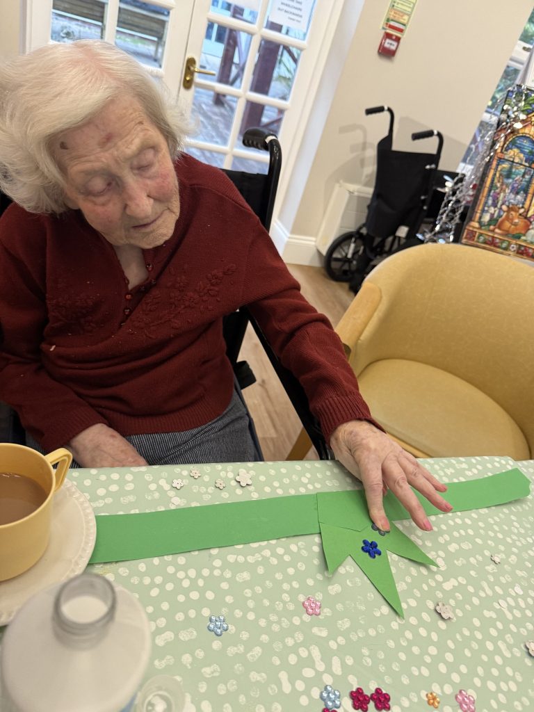 Elderly woman decorating a green paper crown with sequins at a craft table.