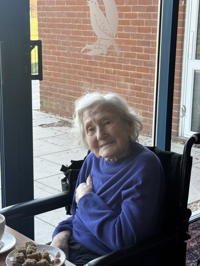 Elderly woman in a wheelchair smiling by a window with a plate of snacks nearby.