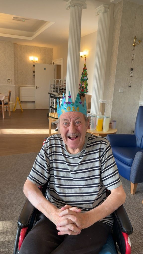 Elderly man wearing a festive crown, smiling while seated in a communal room.