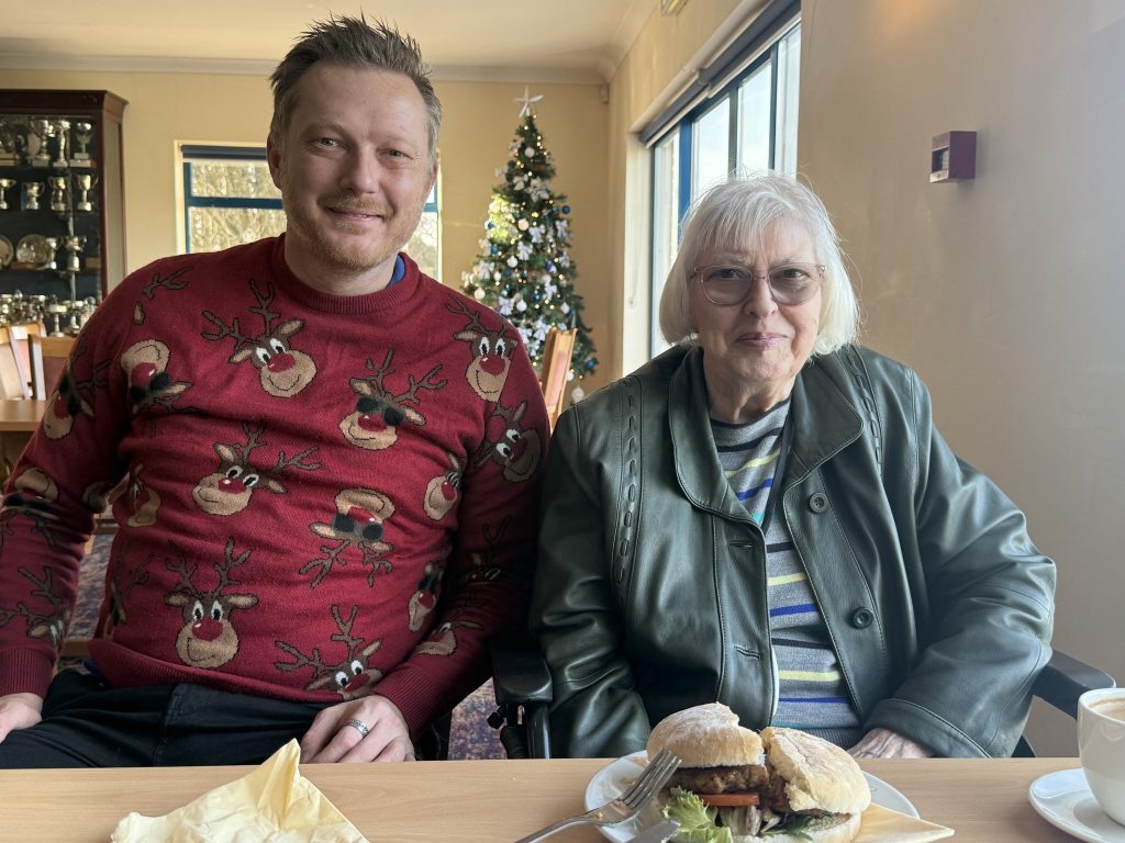 Man and elderly woman sitting together at a table with sandwiches, smiling.
