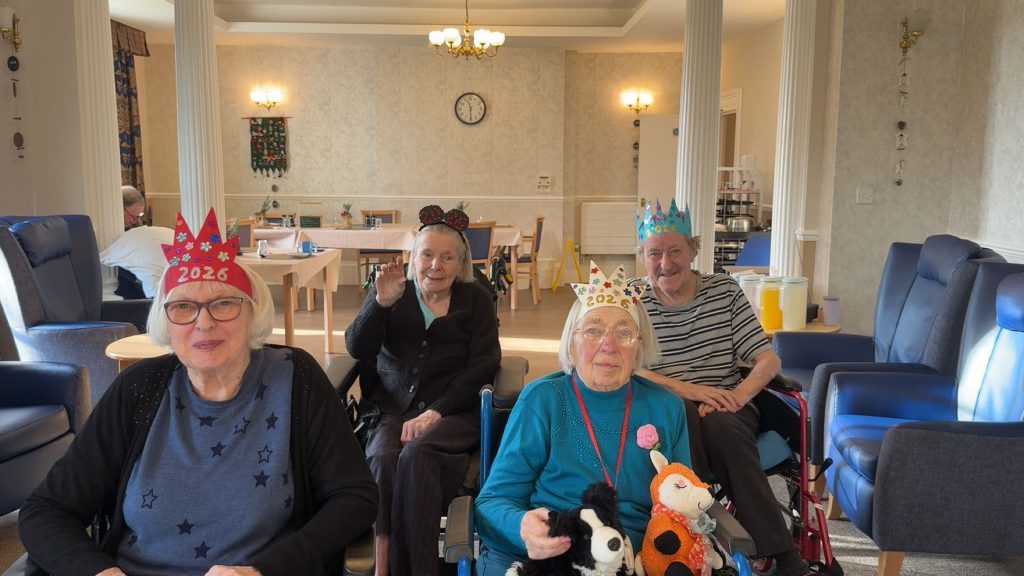 Group of elderly residents wearing colorful crowns, smiling in a bright communal room.