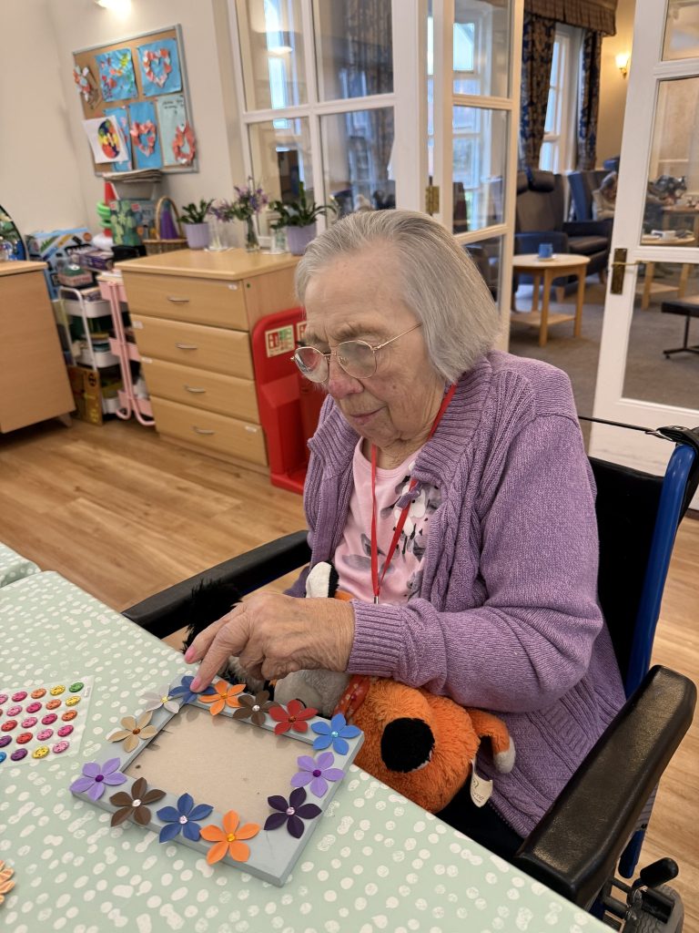 Elderly woman decorating a photo frame with paper flowers while holding a soft toy.